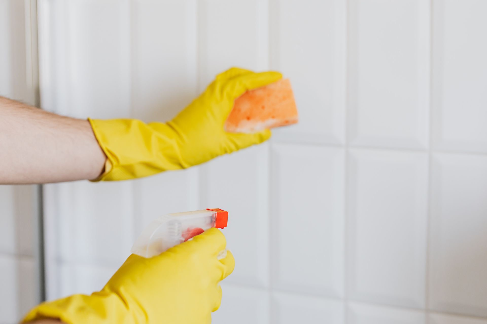 Gloved hands scrubbing bathroom wall tile with a sponge during a deep cleaning